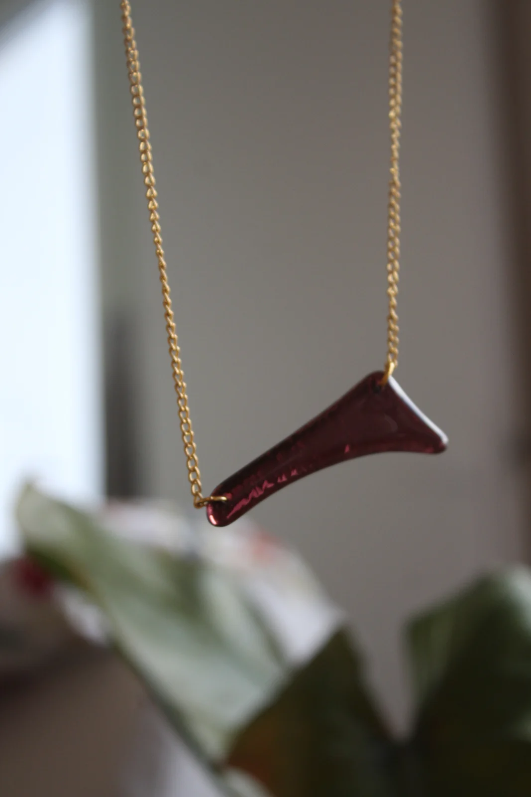 Collier à chaîne dorée avec pendentif asymétrique en verre bordeaux suspendu devant un feuillage