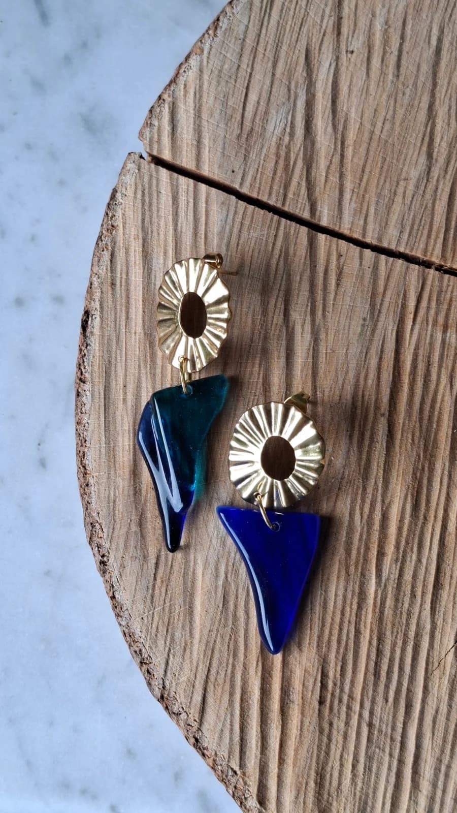 Boucles d'oreilles pendantes dorées avec morceaux de verre bleu et turquoise posées sur un socle en bois