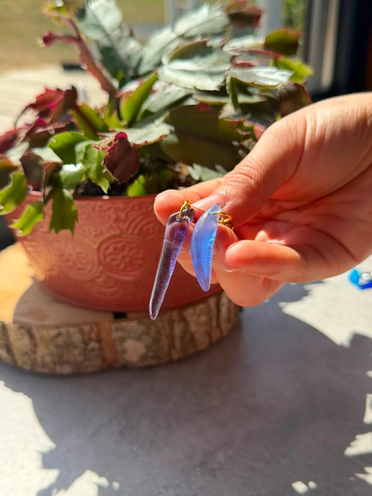 Main tenant deux boucles d’oreilles pendantes en verre translucide violet et bleu à la lumière du soleil.
