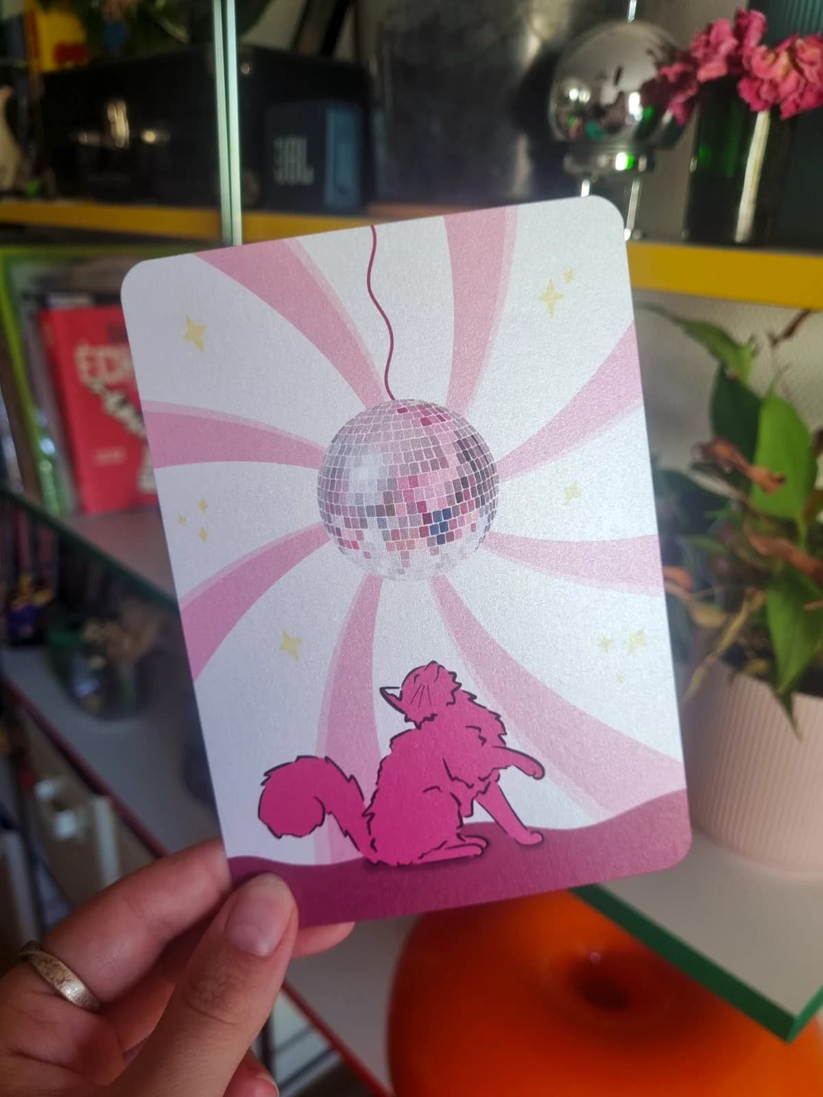 Carte illustrée montrant un chat rose sous une boule à facettes sur fond de rayons roses.