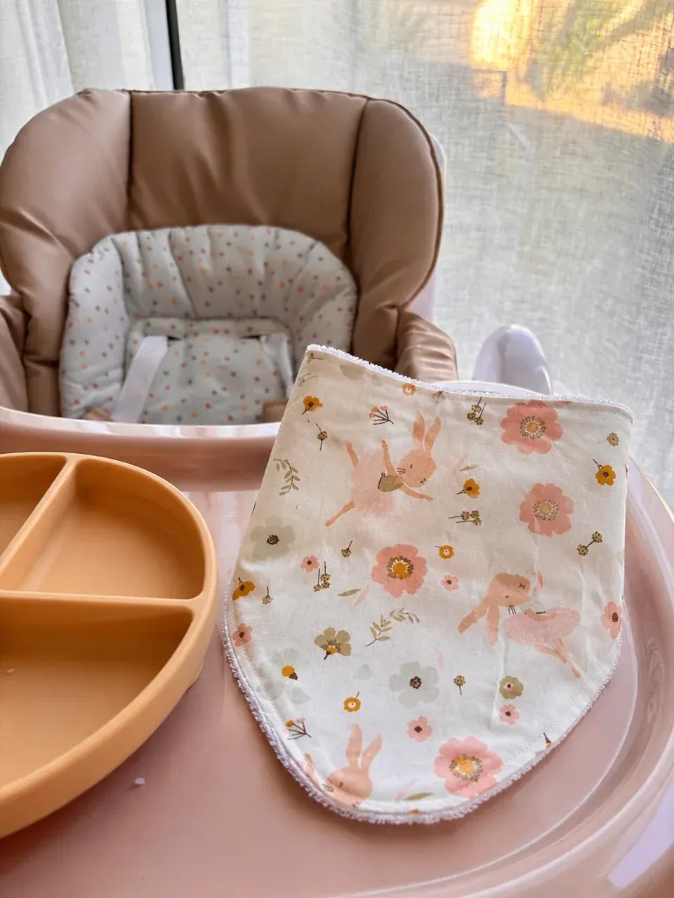 Bavoir bandana à motifs de lapins danseurs et fleurs roses posé sur le plateau d'une chaise haute.
