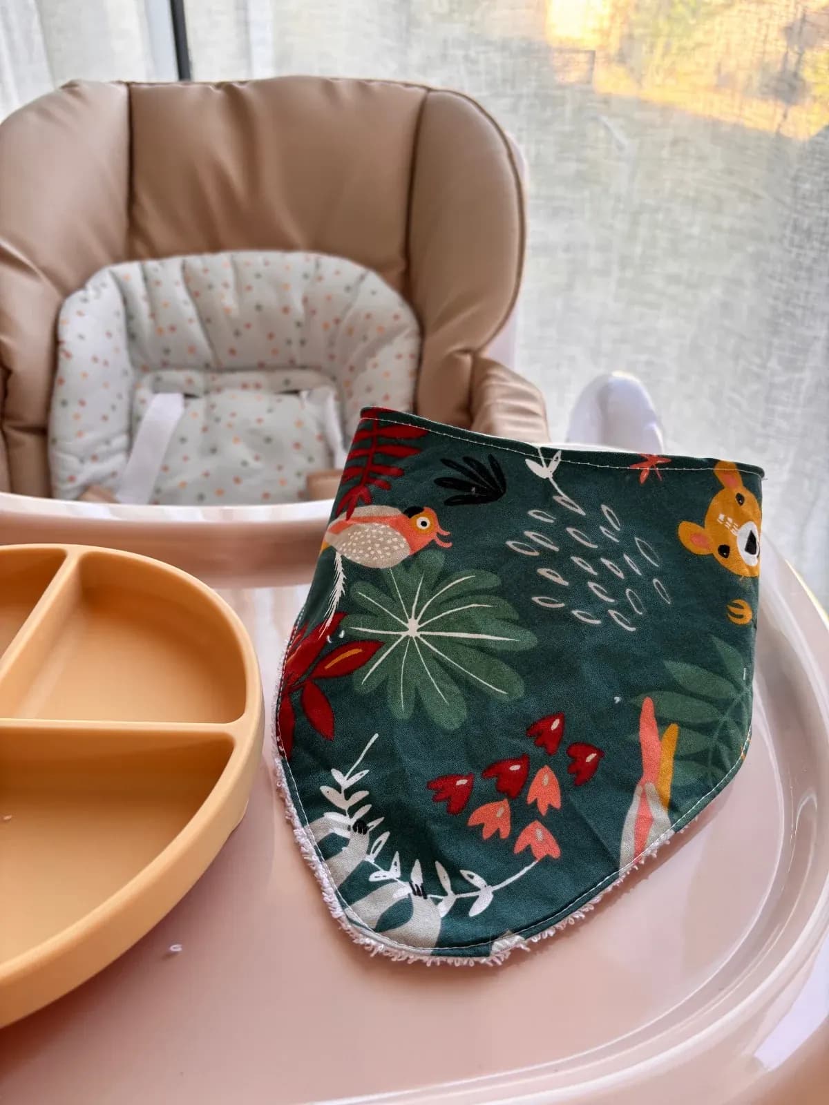 Bavoir bandana vert à motifs animaux et feuillage sur la tablette d'une chaise haute.