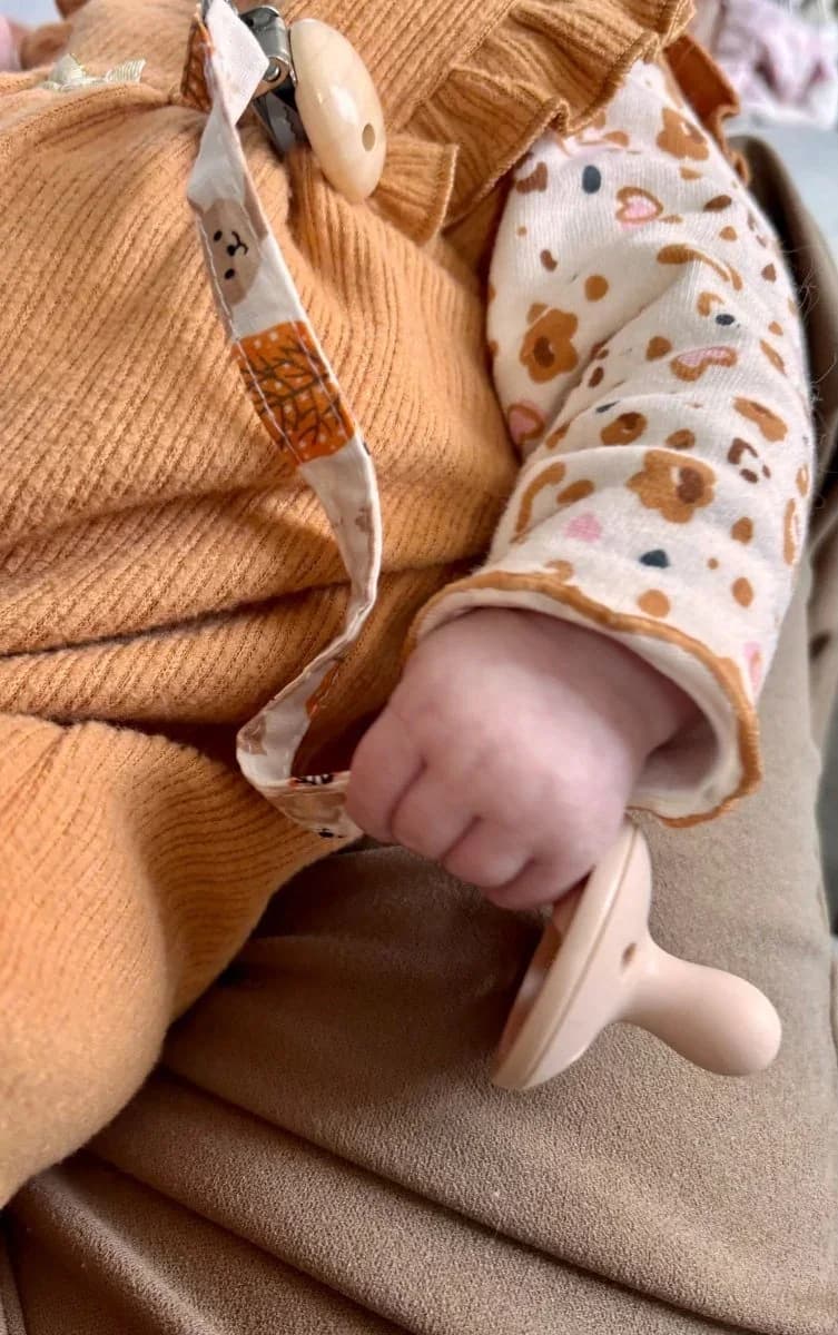 Attache-tétine en tissu à motifs d'automne portée par un bébé en tenue ocre tenant sa tétine.