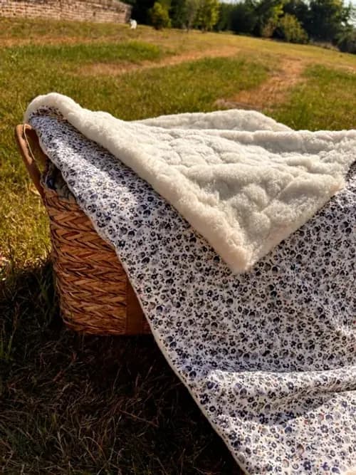 Couverture pour bébé à motif floral bleu et revers polaire blanc, posée sur un panier en osier en extérieur.