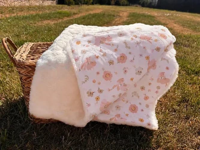 Couverture bébé double-face à motif floral rose et polaire écru, posée sur un panier en osier dans l'herbe.