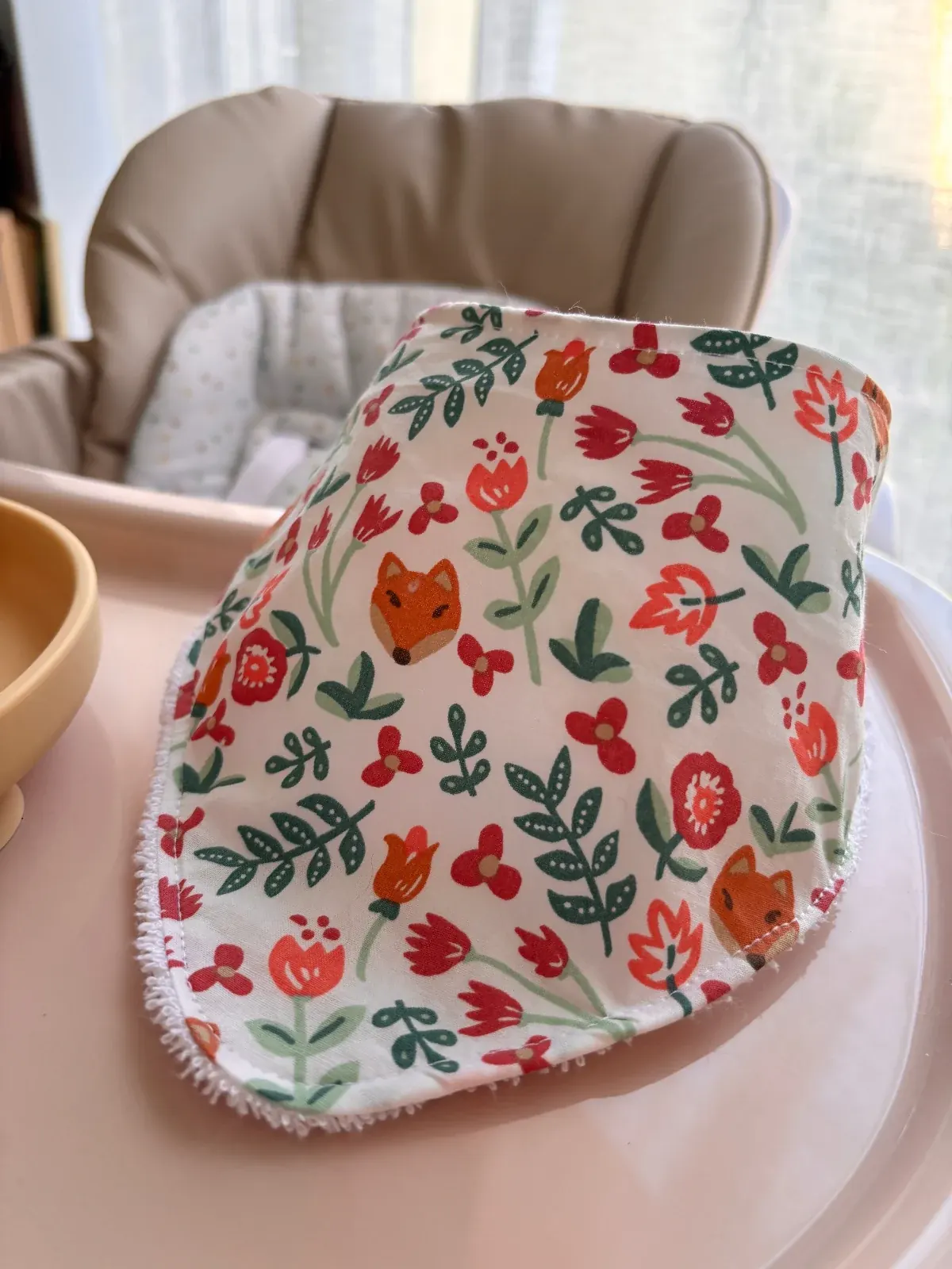 Bavoir bandana déplié à motifs de petits renards et fleurs posé sur une tablette à côté d'une assiette.