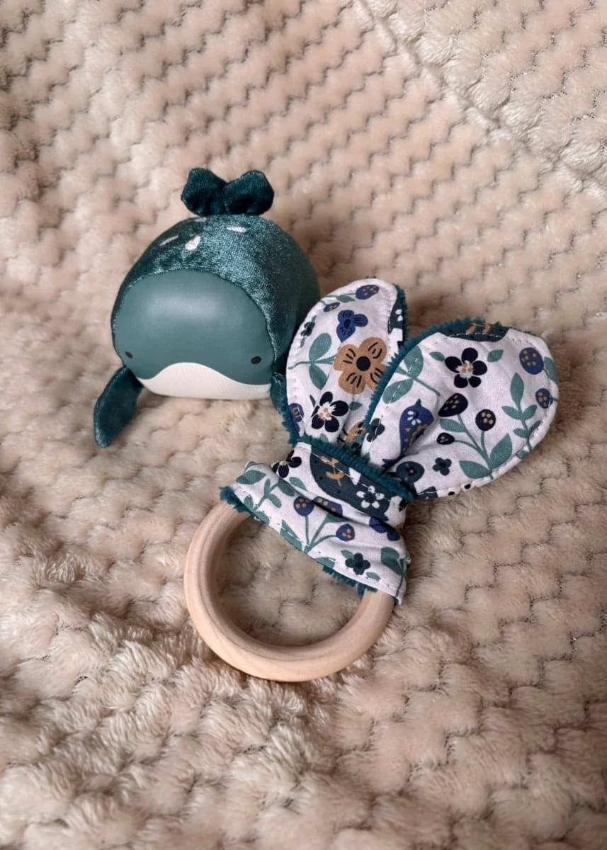 Anneau de dentition en bois avec oreilles en tissu fleuri bleu et vert, posé sur un plaid beige.