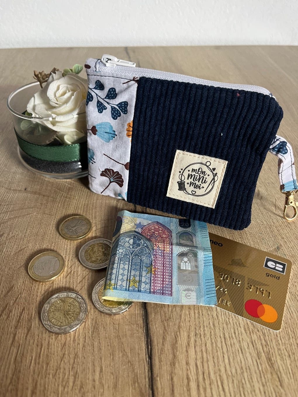 Porte-monnaie zippé bleu côtelé et tissu végétal posé sur une table avec monnaie et carte bancaire.