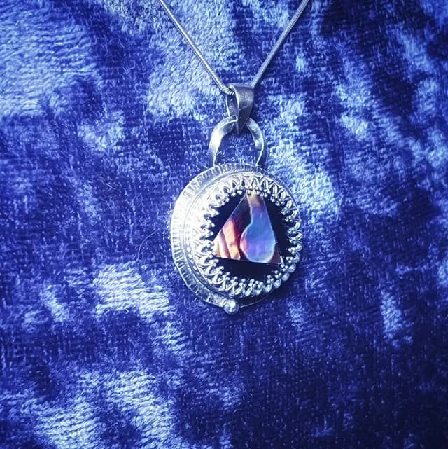 Pendentif circulaire en argent avec centre triangulaire en abalone, posé sur velours bleu.