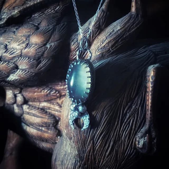 Pendentif artisanal avec pierre ovale grise sertie et croissant de lune sur fond de bois sculpté.