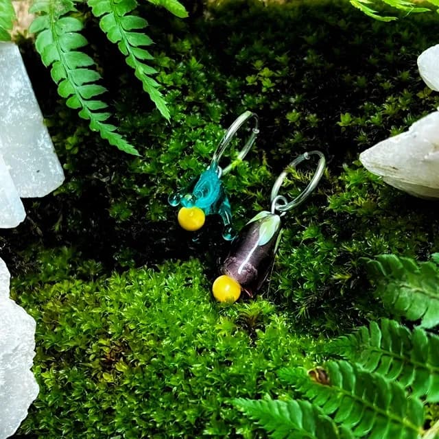 Créoles asymétriques ornées de pendentifs en verre bleu et violet avec billes jaunes, posées sur de la mousse.