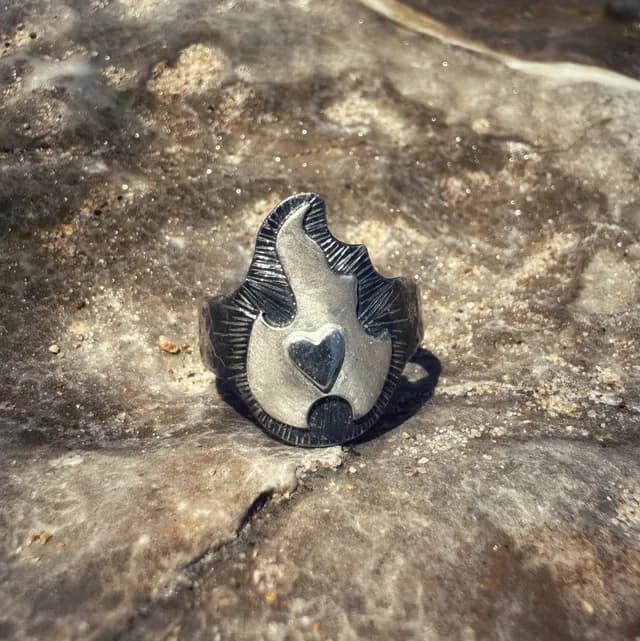 Bague en argent ornée d'un motif de flamme et d'un petit cœur en relief, posée sur une roche.