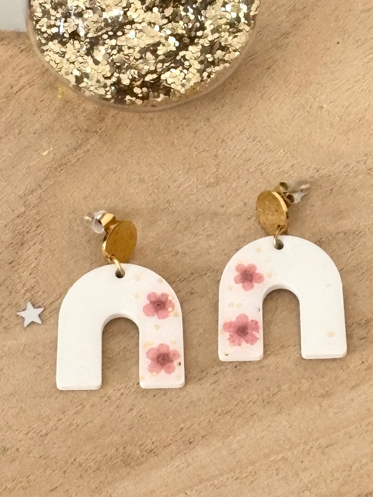 Boucles d'oreilles arc-en-ciel blanches à fleurs roses et paillettes dorées posées sur bois.