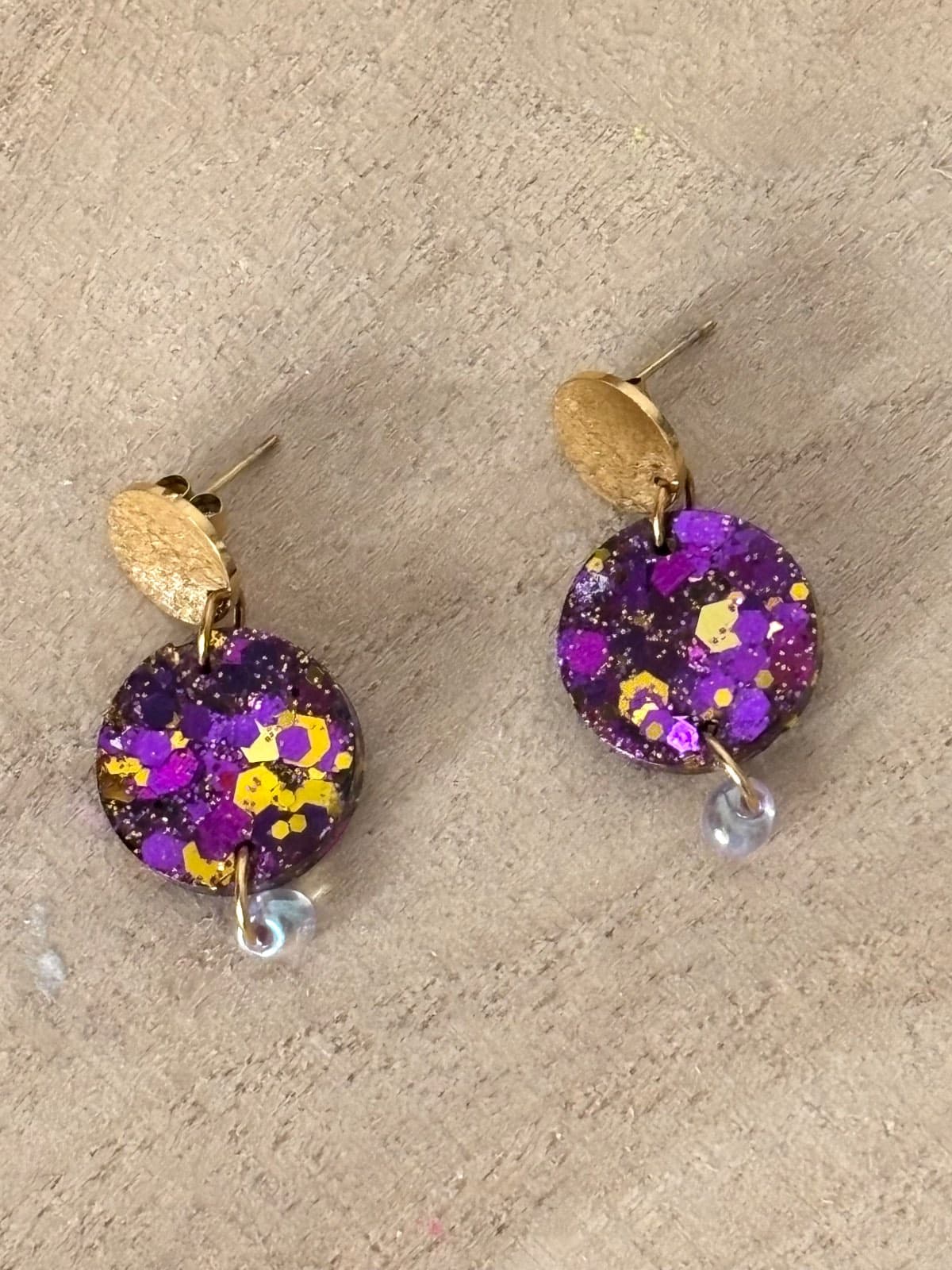 Boucles d'oreilles pendantes rondes violettes et dorées pailletées posées sur du bois.