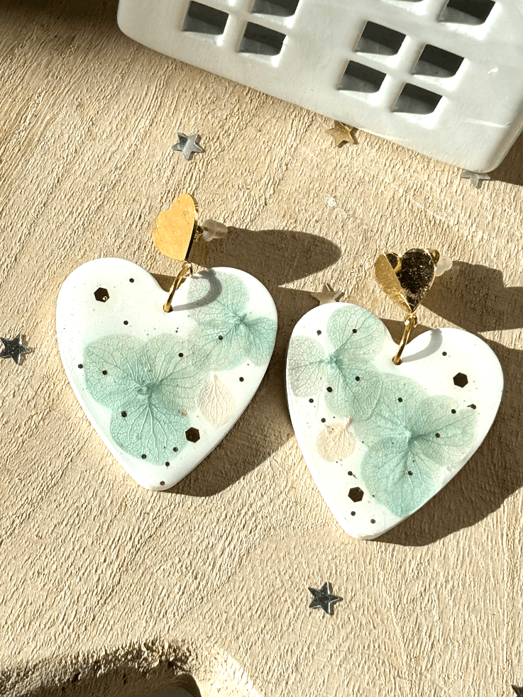 Paire de boucles d'oreilles cœurs blancs à fleurs d'hortensia et attaches dorées sur fond clair.