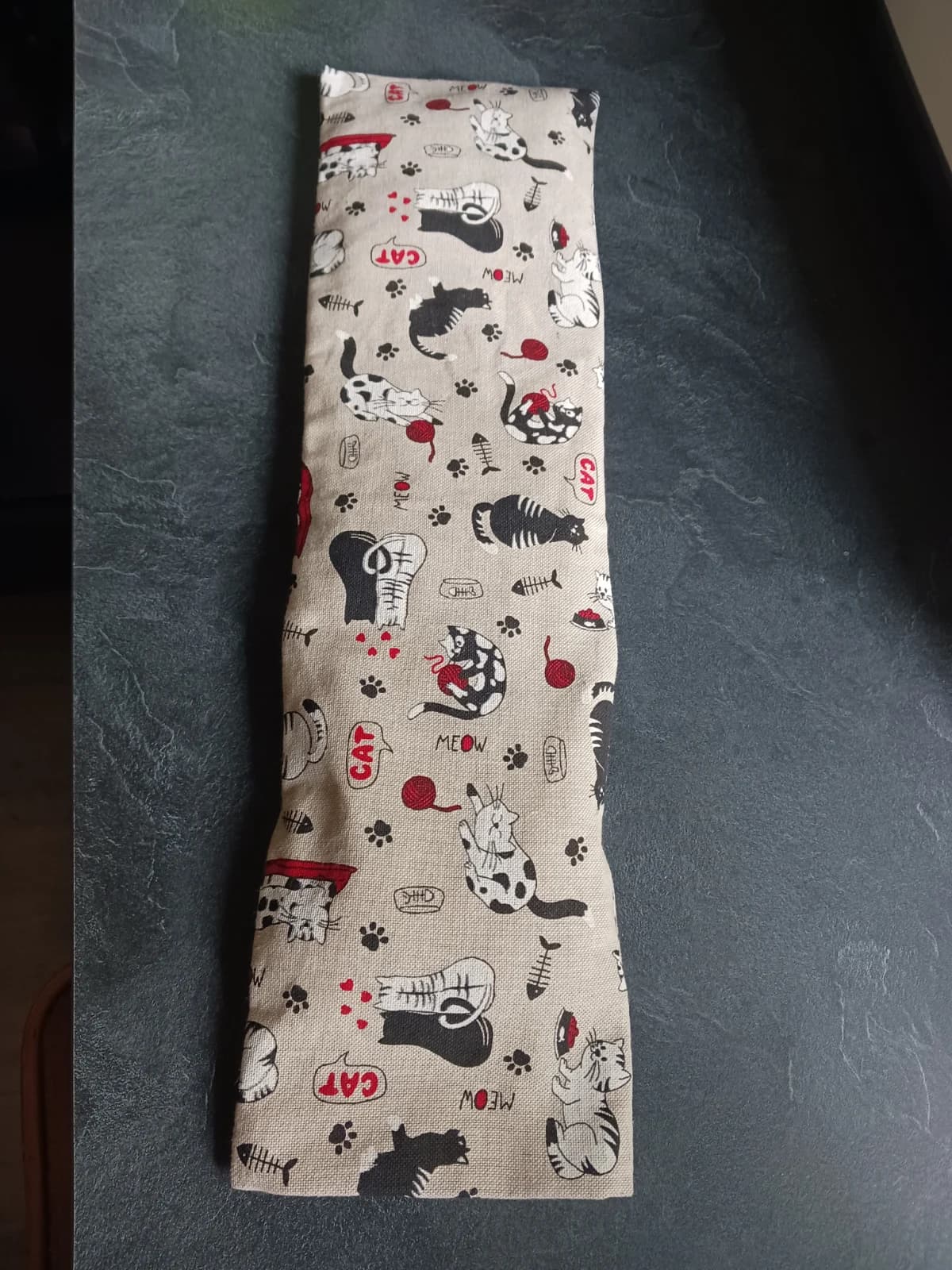 Bouillotte longue en tissu beige à motifs de chats et pelotes rouges posée à plat.