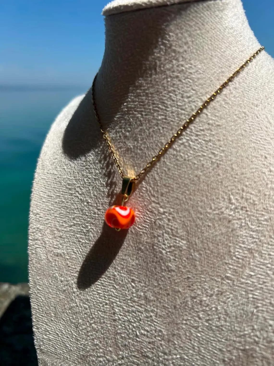 Pendentif perle en verre filé orange sur chaîne dorée porté par un buste d'exposition face à l'eau.