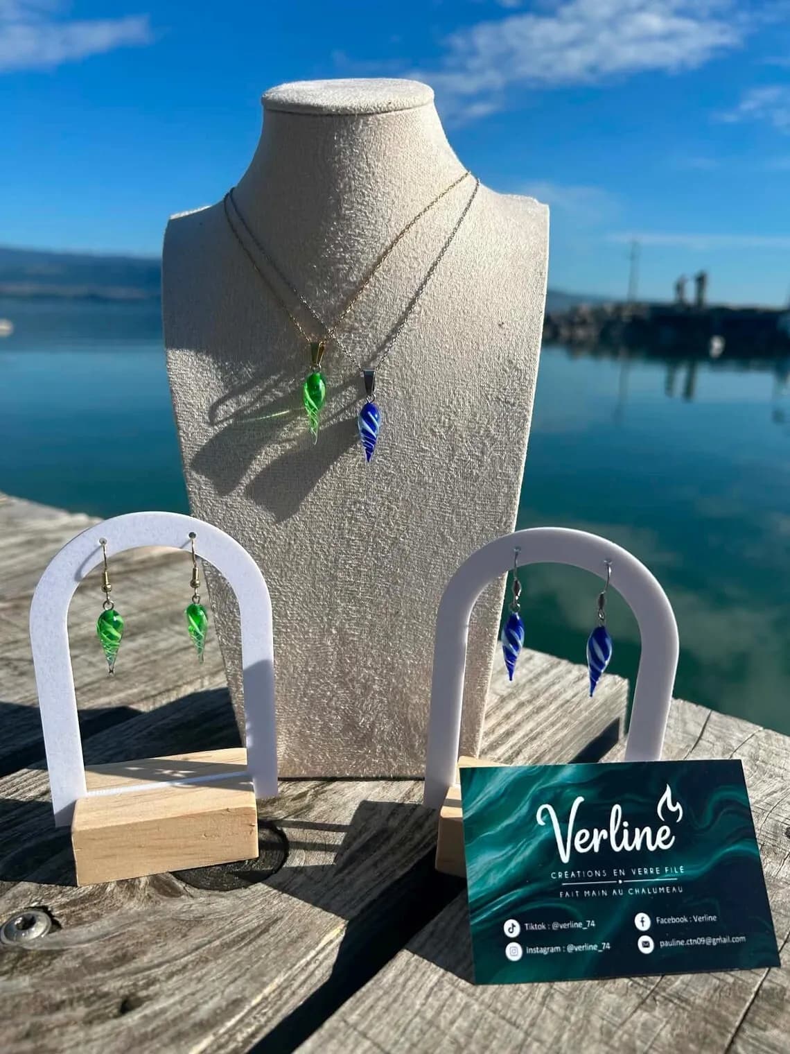 Colliers et boucles d'oreilles pendantes en verre vert et bleu en forme de goutte face à un lac.