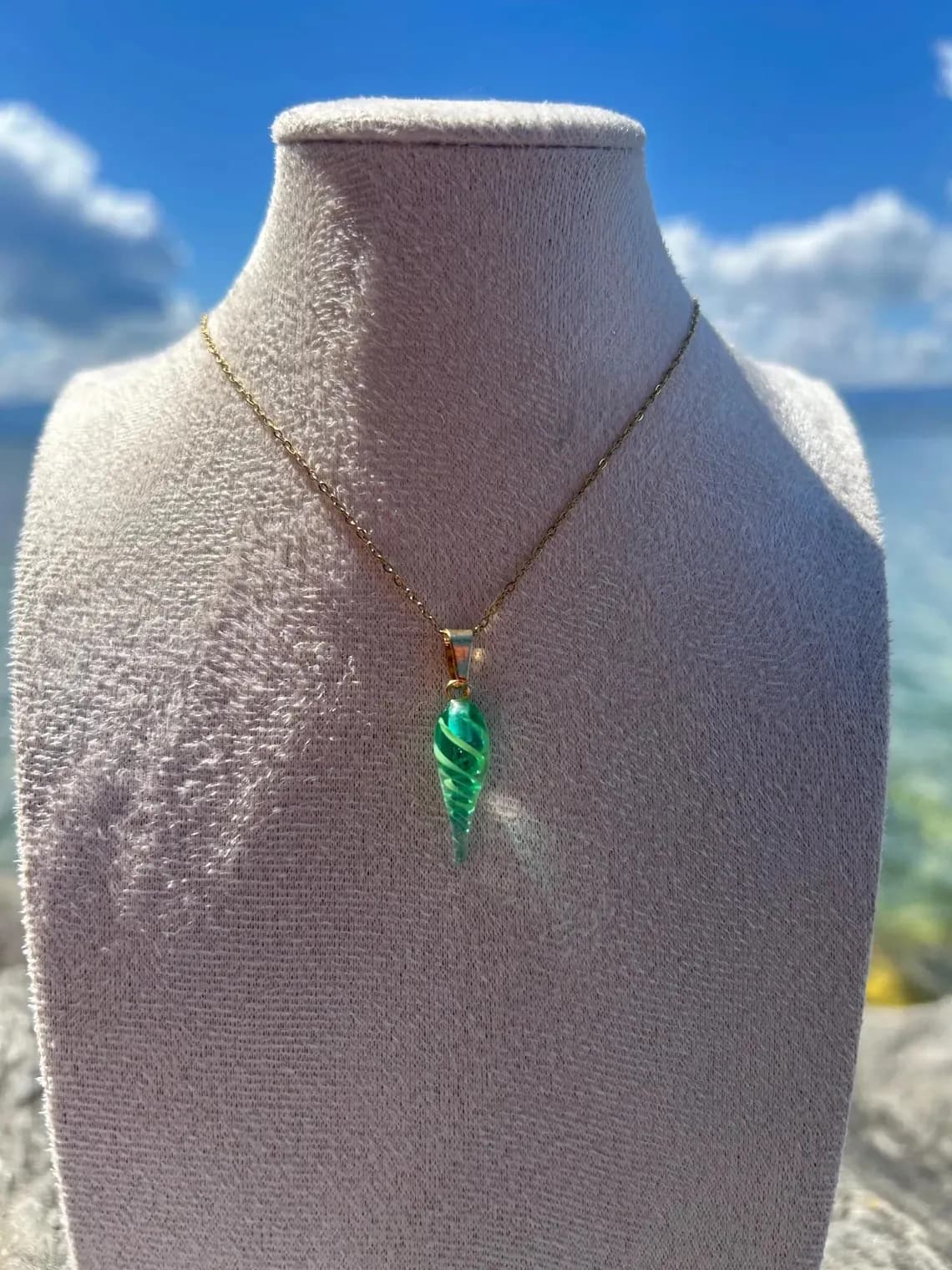 Pendentif coquillage vert en verre sur fine chaîne dorée, présenté sur un buste devant la mer.