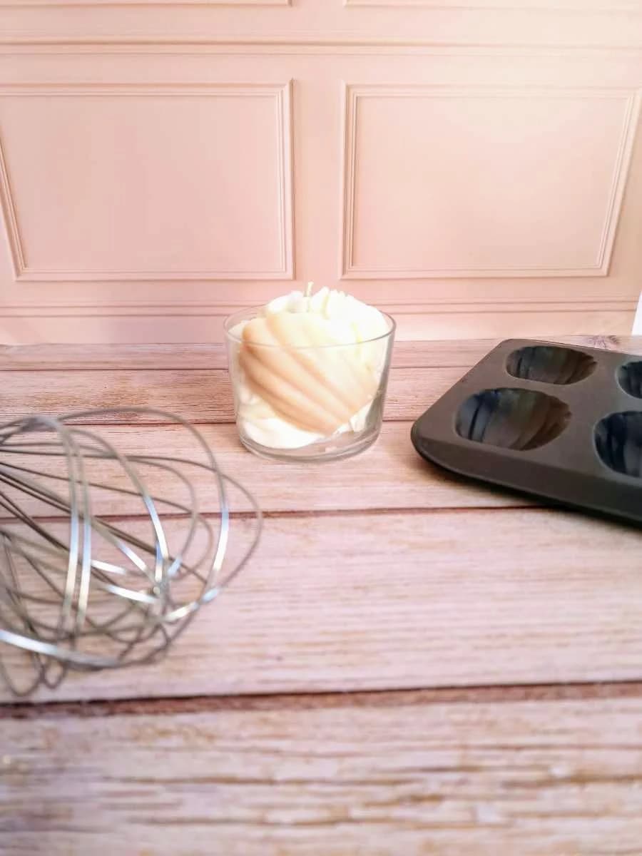 Bougie en verre garnie d'une madeleine en cire beige et crème, posée près d'un fouet et d'un moule